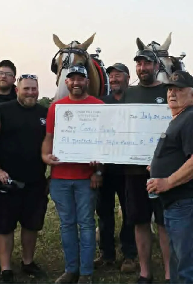 Club members presenting a cheque in a field at dusk, with a tractor and draft horses behind them