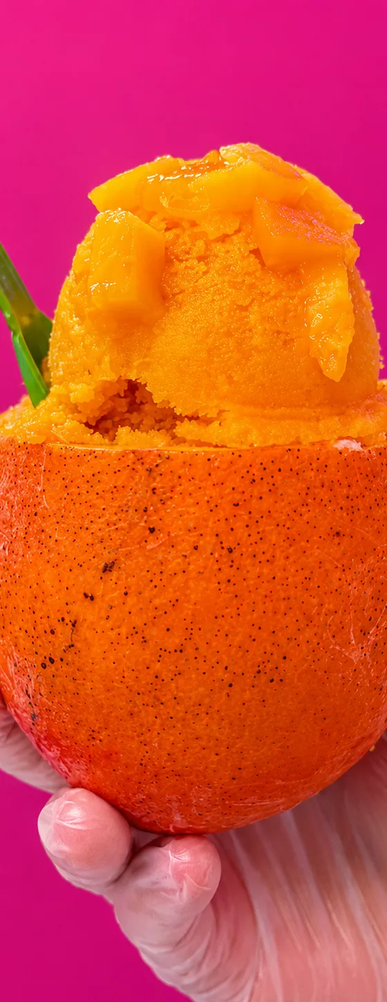 A hand holding a hollowed orange filled with mango sorbet and fresh mango chunks