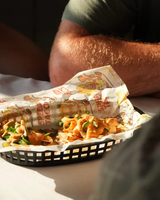 A basket of loaded fries in Speedy Tacos paper on a sunlit table, someone seated behind it
